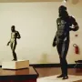 Two Riace bronzes on pedestals in a gallery, with one larger bearded nude male statue in the foreground and a smaller figure behind it.