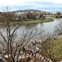 Wide view of the Vistula River with a cityscape, a riverside path full of people, and bare trees in the foreground.