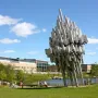 Norra skenet, a large abstract silvery sculpture on a lawn near a pond and modern campus buildings.