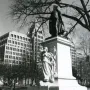 Commodore John Barry statue on a tall pedestal with a relief figure, shown in a park with bare trees and city buildings behind it.