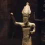 Dark metal statuette of Melqart di Sciacca with a pointed headdress and raised arm, displayed on a light base inside a case.