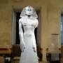 Statue of Amenemhat III standing upright in a museum gallery, wearing a striped nemes headdress and a flared kilt.