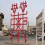 Njé Mo Yé, a bright red abstract sculpture with geometric lines and two stylized figures, stands on a concrete base beside a busy street.