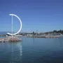 Éole, a large white D-shaped metal sculpture on a rocky breakwater with a marina in the background.