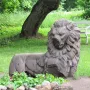 Weathered stone lion sculpture resting on the ground in Mon Repos park, with a tree trunk, bench, and flower bed nearby.