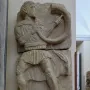 Broken stone relief of the Acrobat from Osuna showing a male figure playing a horn.