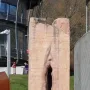 Pi-Chacán, a tall reddish-pink stone sculpture with a deep vertical cleft, standing outdoors near modern buildings and grass.