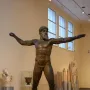 Artemision Bronze, a dark bronze statue of a nude bearded man with outstretched arms in a museum gallery.