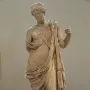 Marble statue of Armed Aphrodite standing in flowing drapery with her head bowed, showing wear and damage.