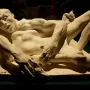 Avarice and Lust by Auguste Rodin, a cream-colored sculpture of two intertwined nude figures against a dark background.