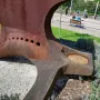 Dorna sculpture with a rusty metal surface, circular holes, and a basin filled with gravel in a park setting.