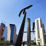 Puerta 1808, a tall angular metallic sculpture, stands in a busy urban plaza with pedestrians, high-rise buildings, and cranes in the background.