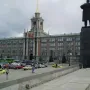 Lenin Monument seen from behind with one arm raised, in front of a large historic building and parked cars.