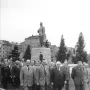 A tall statue of Joseph Stalin stands on a multi-tiered pedestal in a public square, with several men walking in the foreground.