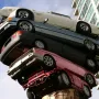 Trans Am Rapture sculpture with stacked cars rising from a large tree trunk against a blue sky, with a modern building in the background.
