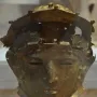 Ribchester Helmet with a patinated bronze face mask and damaged upper section on a display stand.