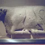 Stone sculpture of the Bull of Osuna in a resting pose on a white display stand.