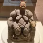 Seated Woman of Çatalhöyük figurine with animal heads on the armrests, displayed in a museum case.