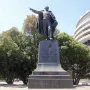 Bronze statue of Benito Juarez standing on a tall inscribed pedestal with trees and a curved building in the background.