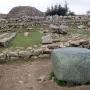 Large greenish stone block in the foreground at Hattusa, with ruins of stone walls and a rocky hill behind it.