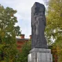 Karl Marx Monument on a tiered stone base with Cyrillic inscriptions, set among trees and a red brick building.