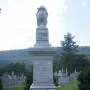 Confederate Memorial stone monument with a draped urn and carved relief in a cemetery, with a small Confederate flag and flowers at its base.