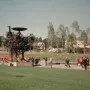Heureka kinetic sculpture standing on a lawn with visitors nearby and trees in the background.