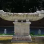 Fontana della Navicella, a weathered stone boat-shaped fountain in an oval basin surrounded by grass and trees.