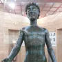 Efebo de Selinunte, a dark bronze nude young man statue with one arm raised in a museum display.