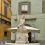 Dolphin Fountain with Triton, a stone sculpture of a bearded man holding a fish in a cobblestone square.