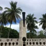 The Torch of Friendship monument’s stone pillar topped with a dark urn and small flame, with palm trees and a low wall behind it.