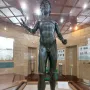 Efebo de Selinunte, a dark bronze nude young man statue with one arm raised in a museum display.