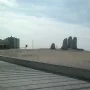 The Hand sculpture with five rounded fingers emerging from a sand dune, with a walkway, cityscape, and ocean in the background.