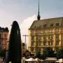 The Brno astronomical clock stands in a sunlit city square with a woman in the foreground and market stalls and buildings behind it.