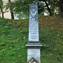 The Pit monument, a tall grey stone memorial with inscriptions and a carved emblem at the top, stands on a grassy hill with wreaths and flower baskets at its base.