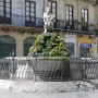 The Fountain of the Genius of Fieravecchia in a city square, with a bearded marble statue on a moss-covered rocky mound behind a protective metal fence.