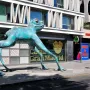 Lucky Frog sculpture with a blue-green oxidized surface and etched symbols on its legs, standing on a city sidewalk.
