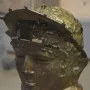 Ribchester Helmet with a patinated bronze face mask and damaged upper section on a display stand.