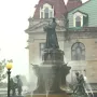 La Fermière bronze statue atop a fountain with water jets, with smaller figures at the base and a historic building behind it.
