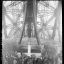 Bust of Gustave Eiffel on a tall pedestal, with floral wreaths and a crowd gathered at the base of the Eiffel Tower.