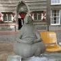 Homunculus Loxodontus sculpture seated on a metal bench outside, with a brick building behind it.