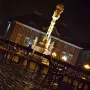 Holy Trinity Monument illuminated at night above wet cobblestones and park benches