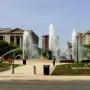 Swann Memorial Fountain in a city plaza, surrounded by paved walkways, trees, and classical stone buildings.