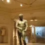Bronze statue of the Athlete from Ephesus standing on a pedestal in a museum gallery.
