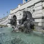 Court of Neptune Fountain with dark metal sea deities and horses rising from a water pool beside an ornate stone building.