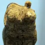 Venus of Hohle Fels figurine with cracked ivory torso, prominent breasts, and a loop at the top on a dark stand.