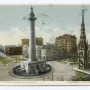 The Washington Monument rises from a public square, with surrounding city buildings and a gothic-style church steeple nearby.
