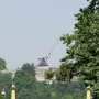 The Prague Metronome, a large red kinetic sculpture, stands on a stone pedestal above a tree-covered hill.