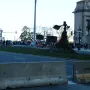 The Bowman statue stands on a stone pedestal beside a monumental archway, overlooking a street with barriers, traffic lights, and people.