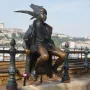 Little Princess bronze statue of a child sitting on a green railing with a city skyline and river in the background.